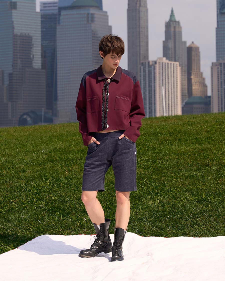 Brando Vassallo Editorial Fashion Lookbook Girl burgundy button down top with denim shoulders and denim shorts and black combat boots. hands in pocket. city skyline in background