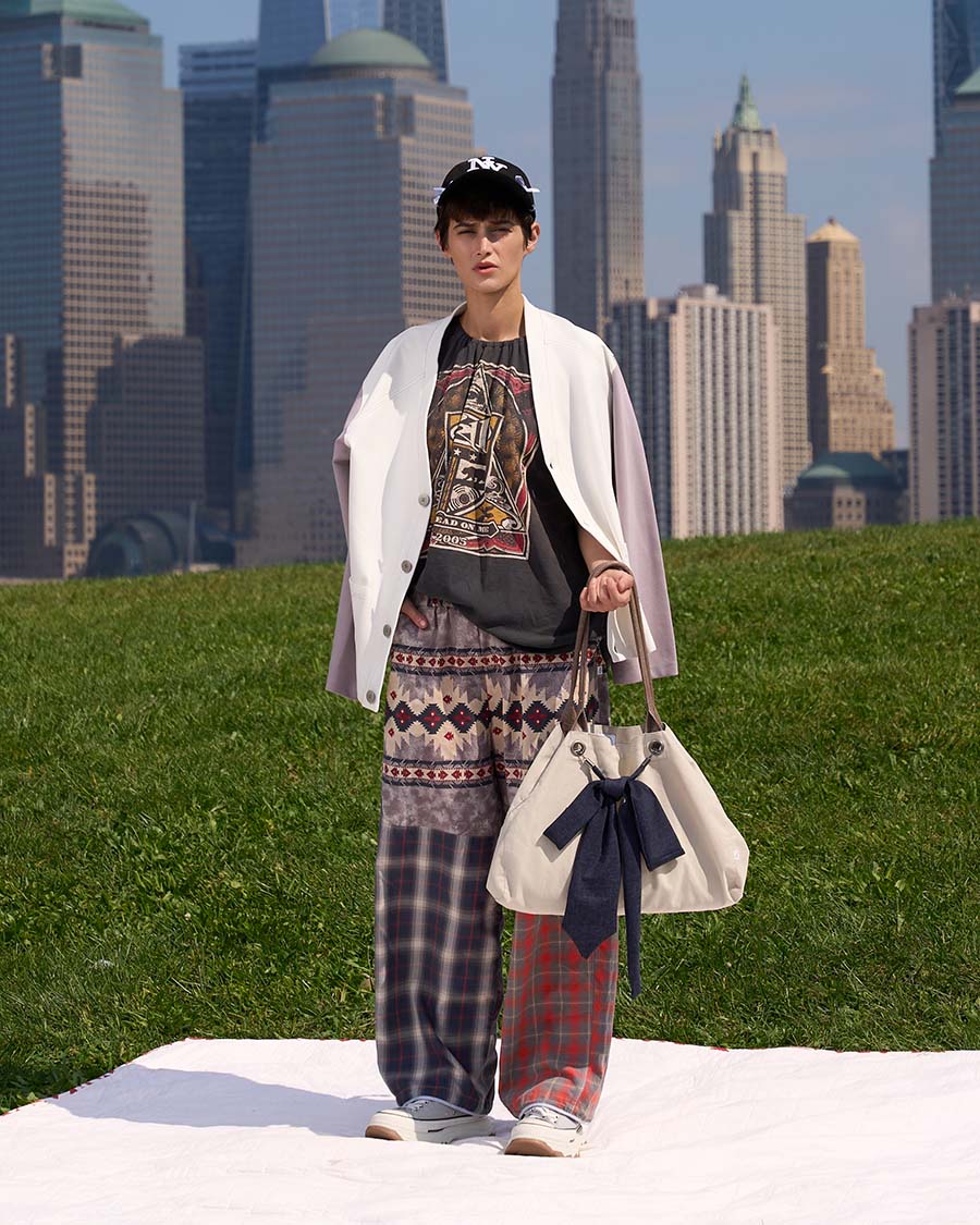 Brando Vassallo Editorial Fashion Lookbook Girl patchwork plaid pants with white cardigan sweater over shoulders holding a tote bag with a dress tie bow and black hat. city skyline in background